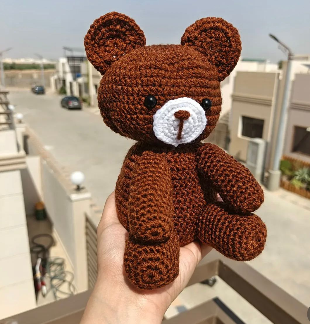 A handheld 8-inch brown Kashmiri handmade crochet teddy bear with a white muzzle and black eyes.